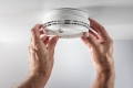 A pair of hands testing a smoke alarm installed on a residential ceiling.