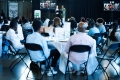 People sitting at tables in a large room as two women speak behind a podium