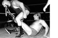 Two wrestlers are in a ring. One is sitting on the mat, while the other stands over him, appearing to bite his shoe. Both wear wrestling attire.