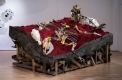 multimedia sculpture made of a wooden rectangular base topped with a stone-like material and a dark red top. Various objects are set on top of the sculpture and resemble horse skeletons.