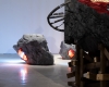 In background, a large rock with lights imbedded into it. In foreground, a portion of a sculpture that resembles stone with a tire rim imbedded into it.