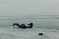 Photograph of a half-human/half-fish figure with long dark hair lying on their stomach on ice at the edge of a body of water. The horizon is visible in the distance.