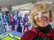 A smiling female volunteer wearing a white mask, blonde wig and red cape/Une bénévole souriante portant un masque blanc, une perruque blonde et une cape rouge.